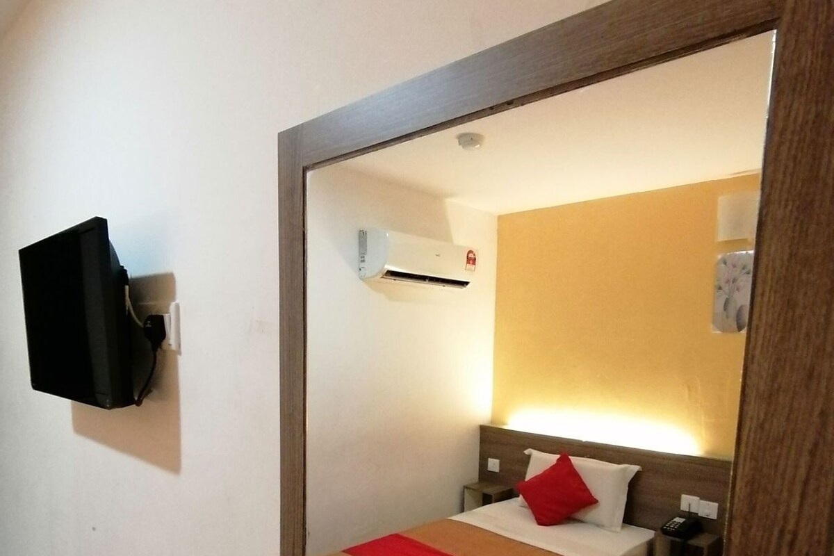 A hotel room with a bed, bedside table, lamp, air conditioning unit, and a television mounted on the wall.