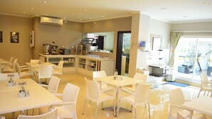 Un comedor de hotel con mesas y sillas blancas, una cocina al fondo y vistas al exterior.