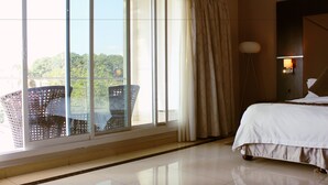 Una habitación de hotel con un amplio ventanal corredero que da a un balcón, una cama con ropa blanca, una lámpara de pie y vistas a zonas verdes.