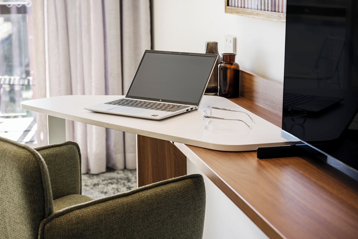 Ein Laptop auf einem weißen Schreibtisch, daneben eine Brille, ein dunkler Bildschirmfernseher und eine braune Vase.