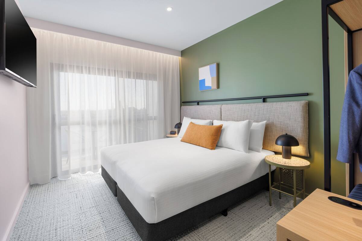 Ein modernes Hotelzimmer mit einem großen Bett, einem Nachttisch mit Lampe, einem an der Wand befestigten Fernseher und Blick auf die Stadt durch transparente Vorhänge.