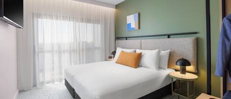 A modern hotel room with a large bed, a bedside table with a lamp, a TV mounted on the wall, and a view of the city through sheer curtains.