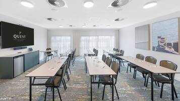 Un salón de conferencias con una pantalla plana, varias mesas, sillas y un amplio ventanal con cortinas.
