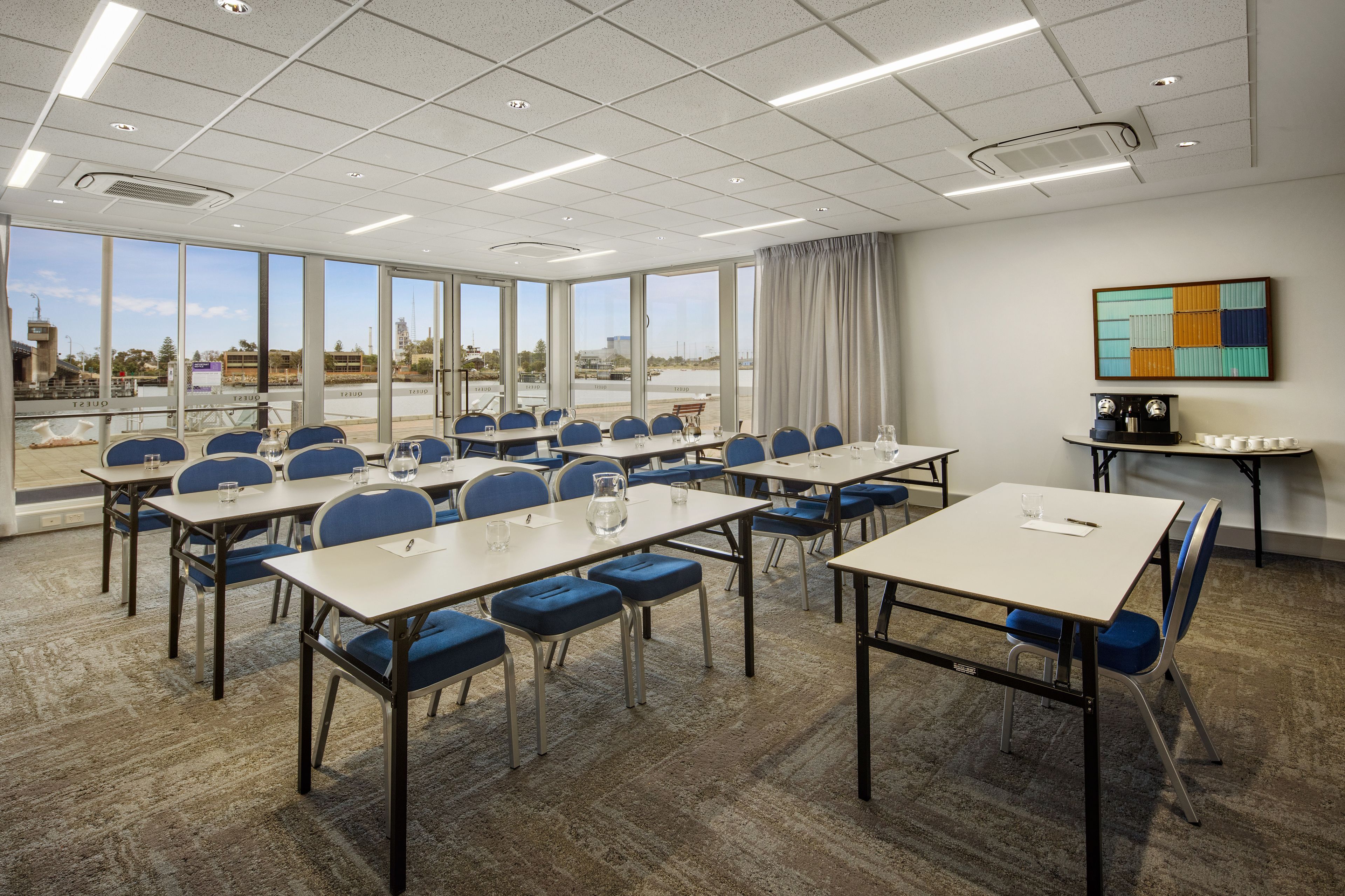 Une salle de conférence dotée de plusieurs tables et chaises, de grandes fenêtres offrant une vue sur l’extérieur et d’un plafond moderne agrémenté de luminaires.