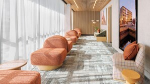 Un hall d’hôtel moderne avec une rangée de fauteuils orange, un tapis à motifs et une grande fenêtre donnant sur un immeuble.