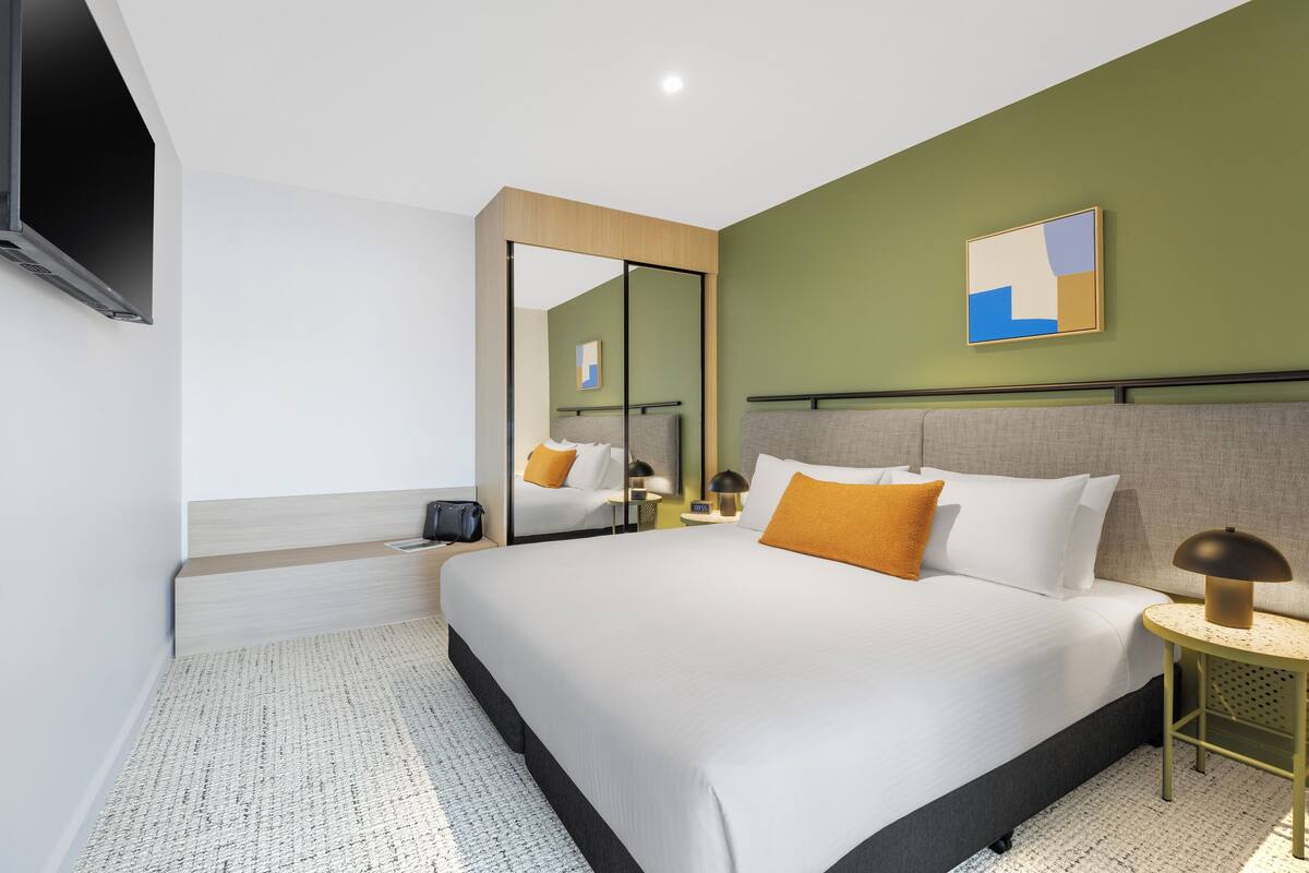Ein modernes Hotelzimmer mit einem großen Bett, einem Nachttisch mit Lampe, einem Spiegel und einem an der Wand befestigten Fernseher.