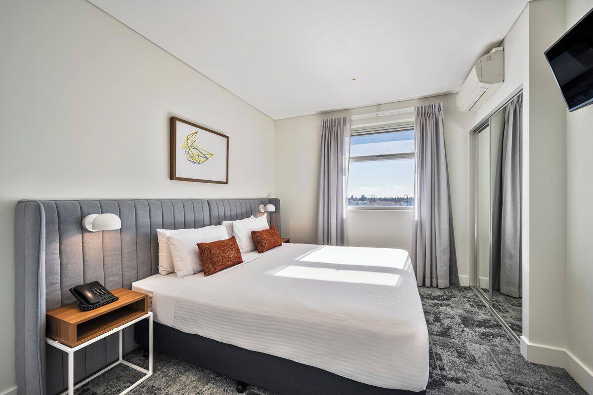 Ein Hotelzimmer mit einem Bett, einem Nachttisch mit Telefon, einem an der Wand befestigten Fernseher und einem Fenster mit Vorhängen.