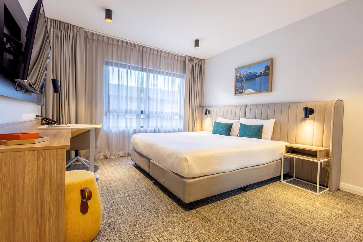 A modern hotel room with a large bed, a desk, a chair, and a painting on the wall.