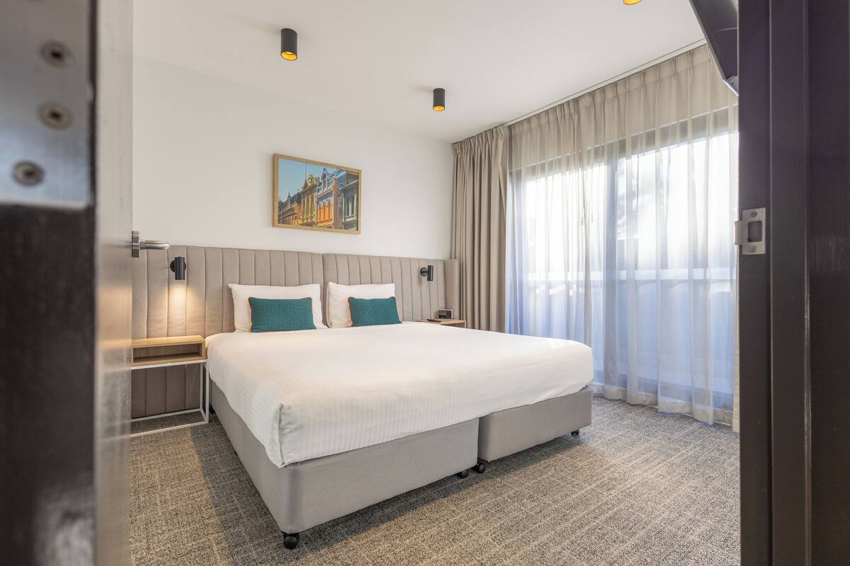 A hotel room with a large bed, a nightstand, a painting on the wall, and sheer curtains on the window.