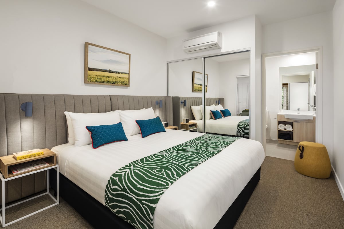 Una habitación de hotel moderna con una cama grande, una mesita de noche con una revista, un espejo, un taburete pequeño y un baño visible a través de una puerta abierta.