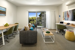 Una sala de estar moderna con un sofá, una mesa de comedor con sillas, una mesita de centro, una computadora portátil sobre un escritorio y un ventanal con vistas a árboles y un edificio.