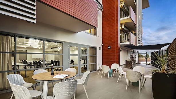 An outdoor dining area with round tables and white perforated chairs, a large potted plant, and a view of the building's interior through glass doors. - Quest Griffith (Griffith)