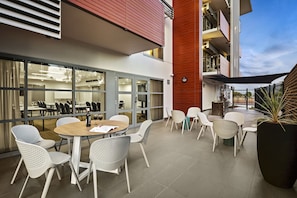 Un área de comedor al aire libre con mesas redondas y sillas blancas perforadas, una planta grande en maceta y vistas del interior del edificio a través de puertas de vidrio.