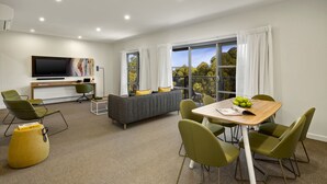A modern living room with a sofa, green chairs, a wooden table with a bowl of fruit, a flat-screen TV, and a painting on the wall. - Quest Griffith (Griffith)