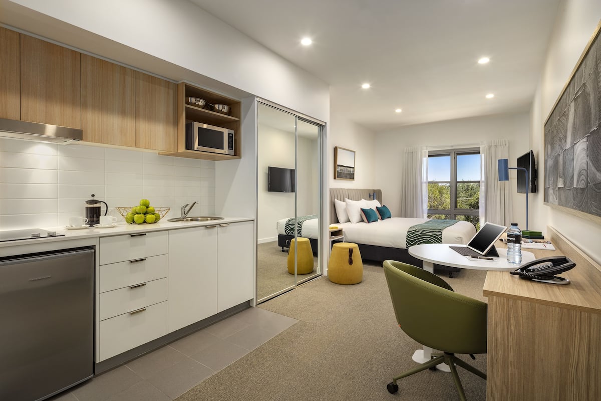 Una habitación de hotel moderna con área de cocina, cama con vistas, escritorio con computadora portátil y mesita de noche con teléfono y botella de agua.