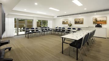 Una sala de conferencias con una larga mesa, sillas y una pizarra. Hay ventanas con persianas, una puerta y un reloj en la pared.