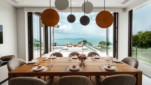 Comedor con una mesa de madera dispuesta para seis comensales, ventanas amplias con vista al mar y lámparas colgantes.