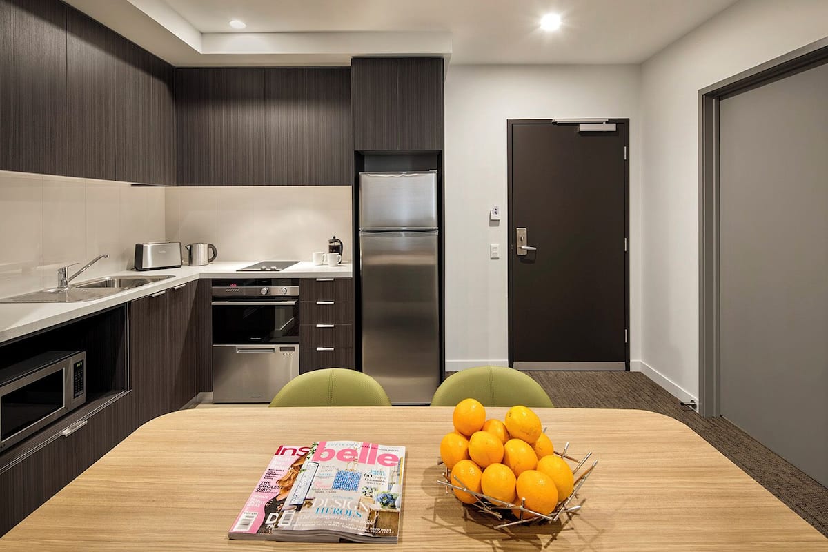Una cocina moderna con armarios de madera oscura, electrodomésticos de acero inoxidable y una mesa de comedor de madera con una revista y un bol de naranjas.