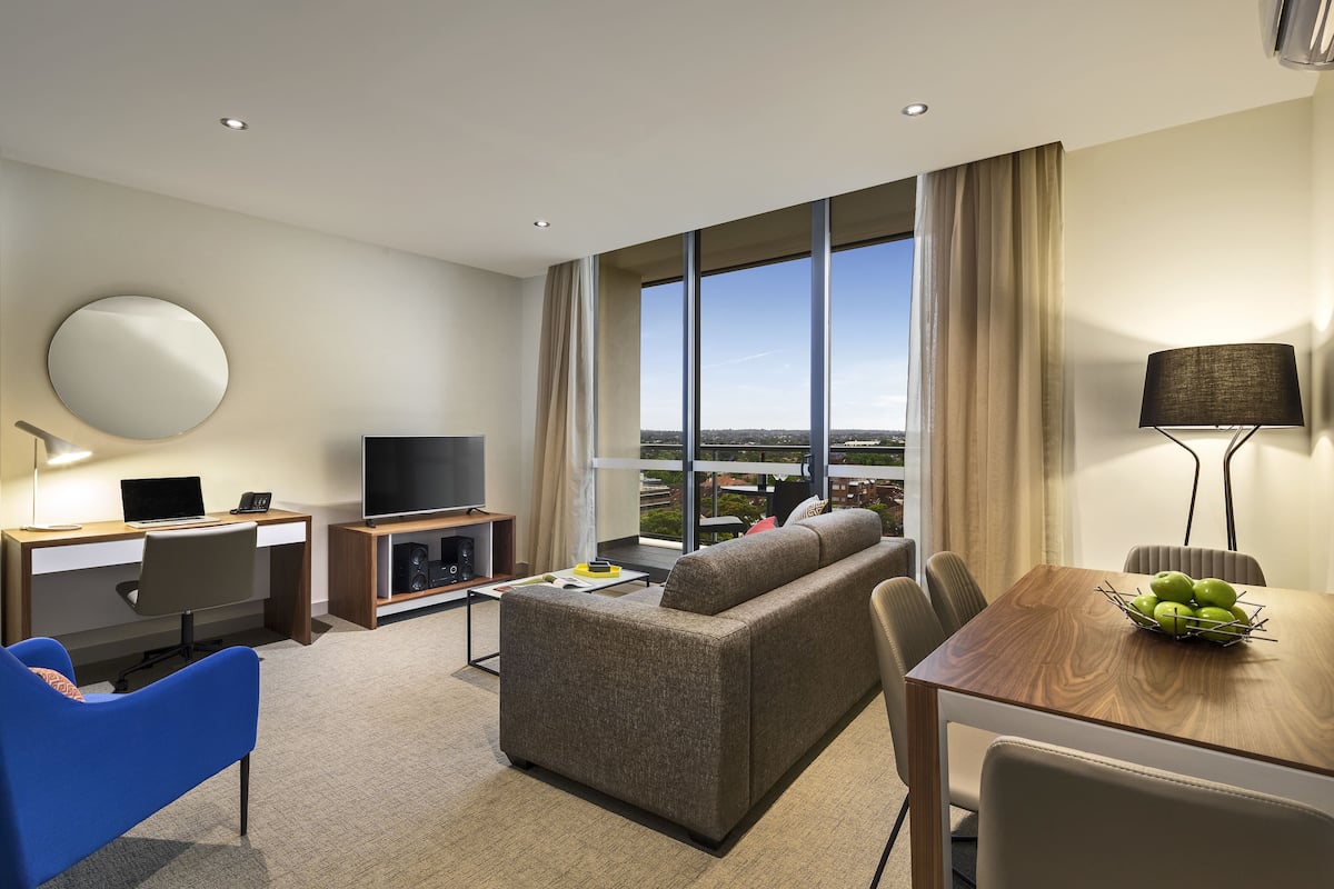 Una habitación de hotel moderna con sofá, mesa de comedor, televisión y un ventanal con vistas a la ciudad.
