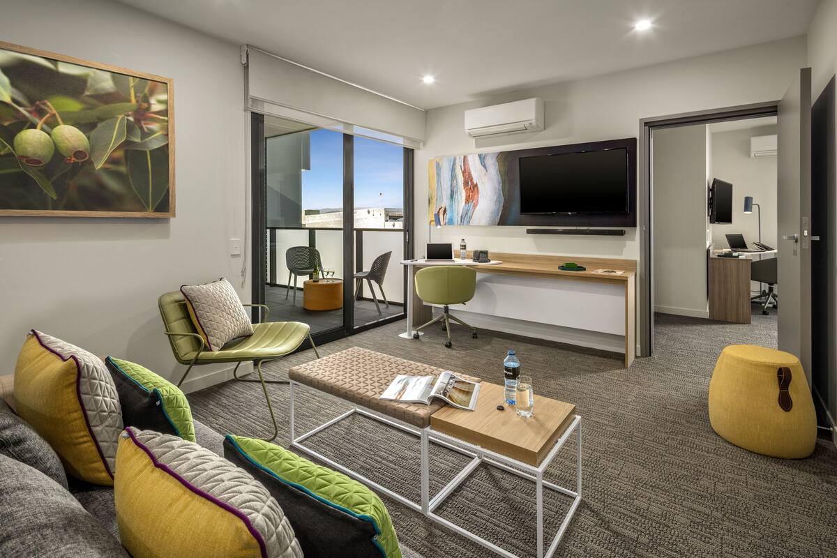 Una habitación de hotel moderna con sofá, una mesa de centro, un escritorio con una computadora portátil, un televisor y vistas al exterior.