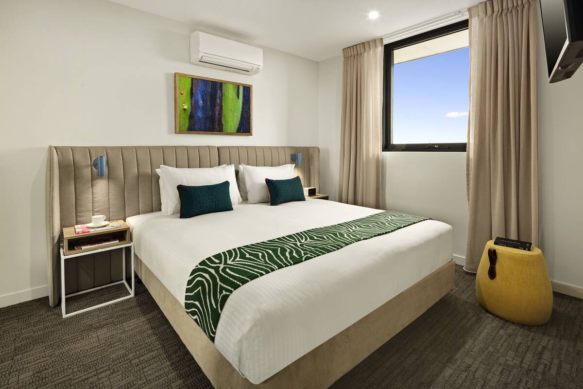 Habitación de hotel con una cama grande, una mesita de noche, una pequeña mesita auxiliar, un aire acondicionado instalado en la pared, una ventana con cortinas y un cuadro en la pared.