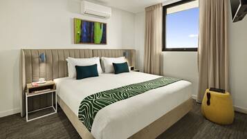 A hotel room with a large bed, a bedside table, a small side table, a wall-mounted air conditioner, a window with curtains, and a painting on the wall.