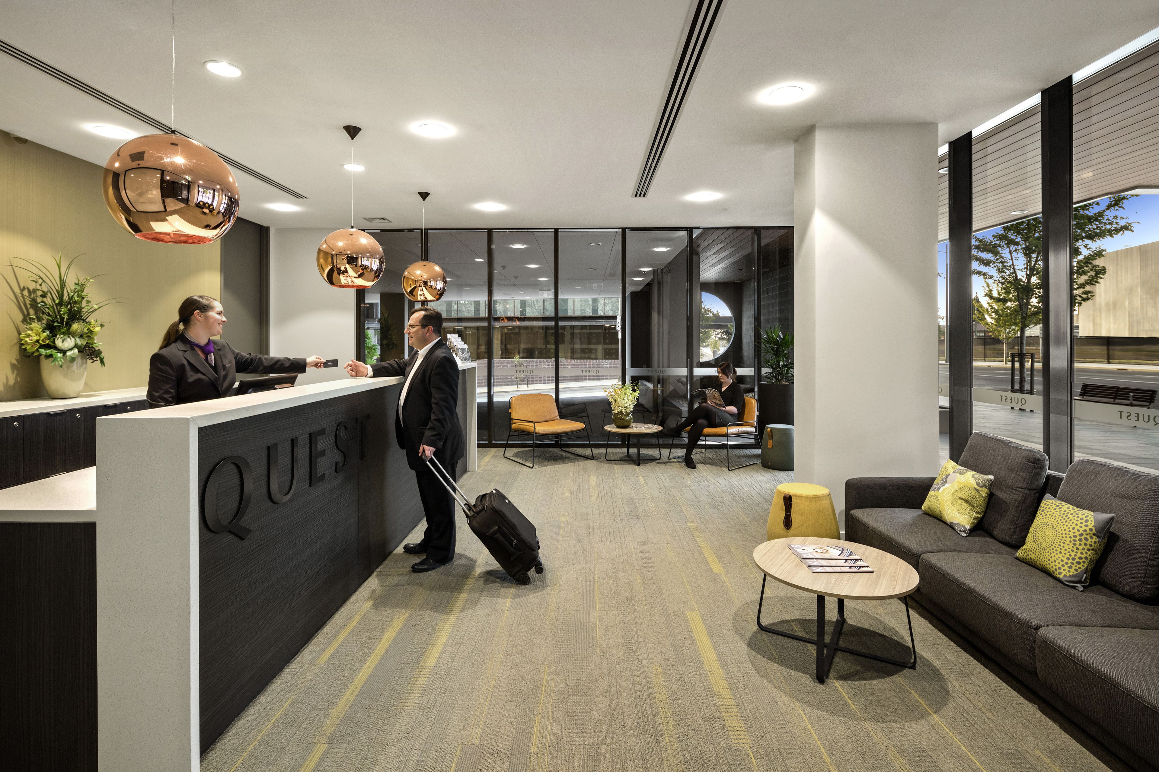 Ein Hotel-Rezeptionsbereich mit modernem Design, mit einer Rezeption mit der Aufschrift "QUEST", einer Sitzecke mit Sofas und Sesseln sowie großen Fenstern mit Blick ins Freie.