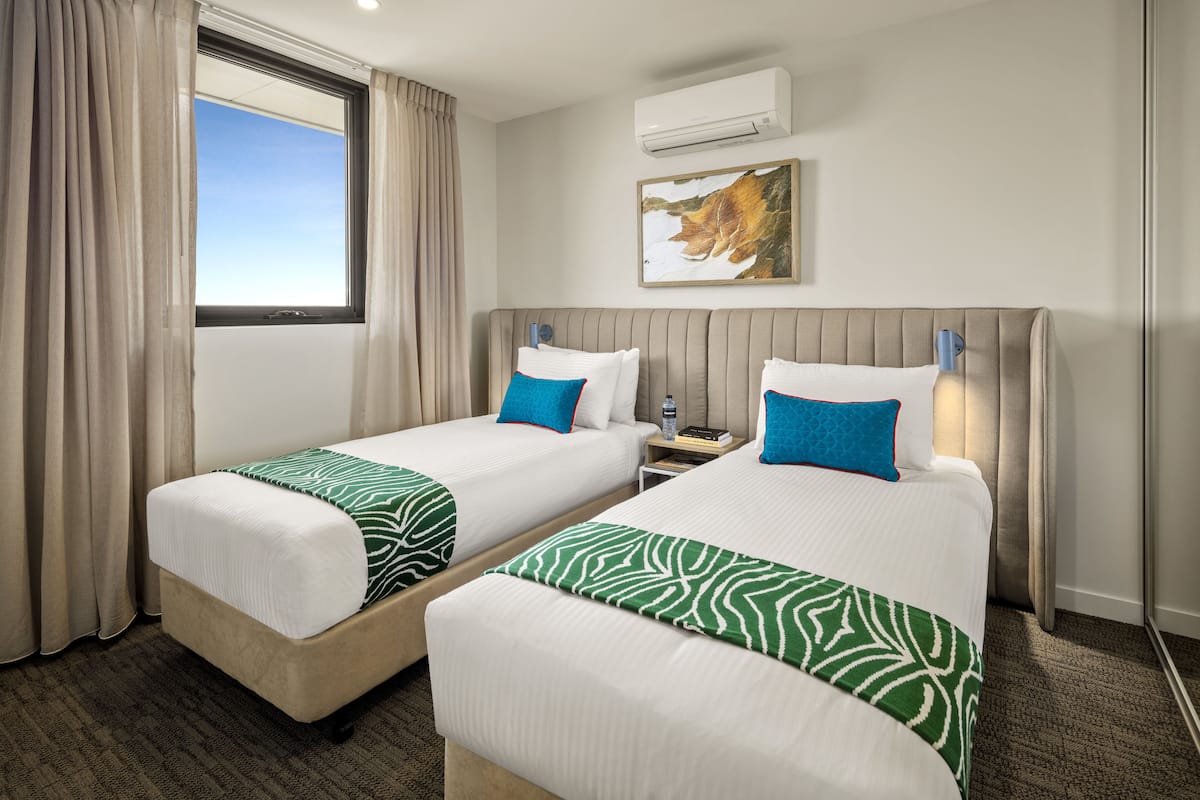 Habitación de hotel con dos camas, cabeceros color beige, ropa de cama blanca, almohadas decorativas en tonos verde y azul, una mesita de noche con lámpara, un cuadro enmarcado en la pared y un equipo de aire acondicionado.