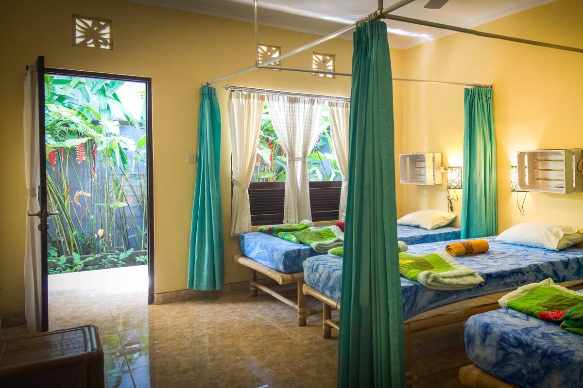 Una habitación con tres camas, ventilador de techo y vista a un jardín por la ventana.