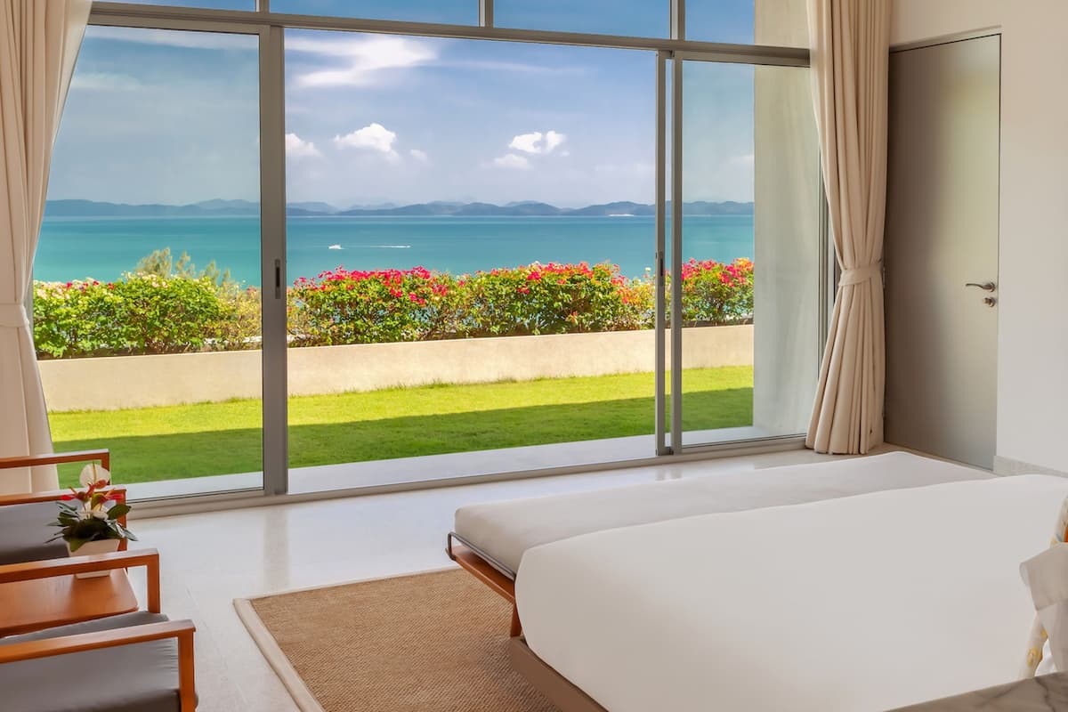 A bedroom with a large window, a bed, a chair, and a view of the sea and mountains.
