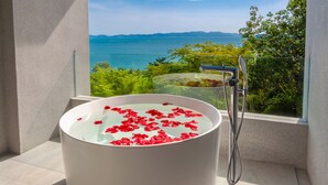 Una bañera llena de agua y pétalos de rosa, con un cabezal de ducha y vistas al mar y a las montañas a través de una ventana.