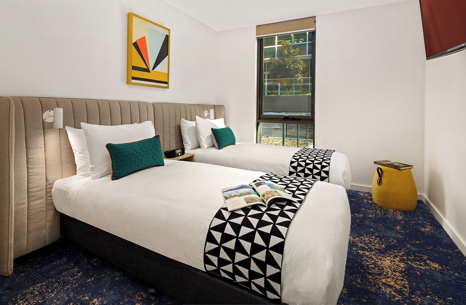 A hotel room with two beds, a large window, a painting on the wall, and a small yellow side table.