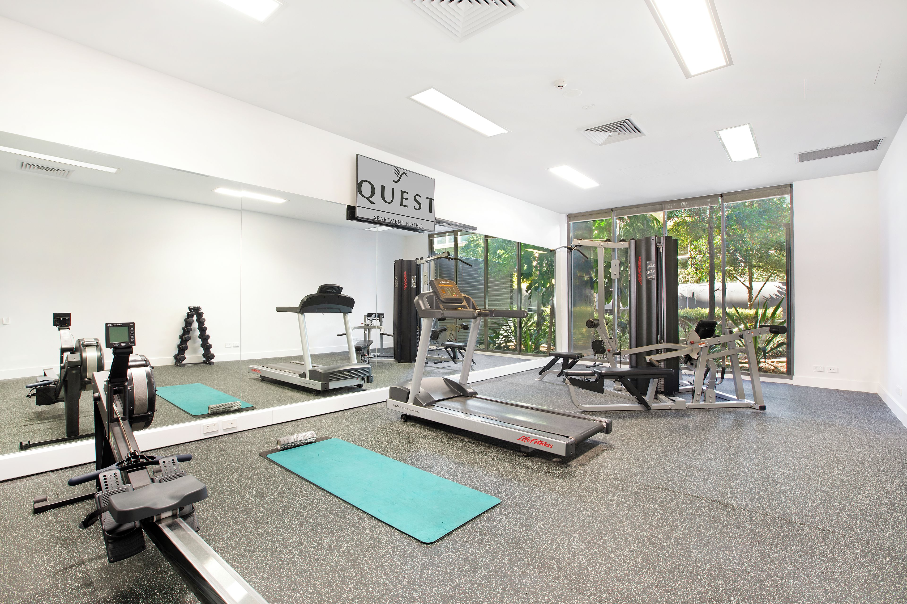 A gym with various exercise equipment, a large mirror, and a sign that reads 'QUEST APARTMENT HOTELS'.