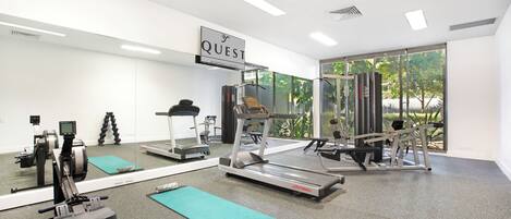 A gym with various exercise equipment, a large mirror, and a sign that reads 'QUEST APARTMENT HOTELS'.