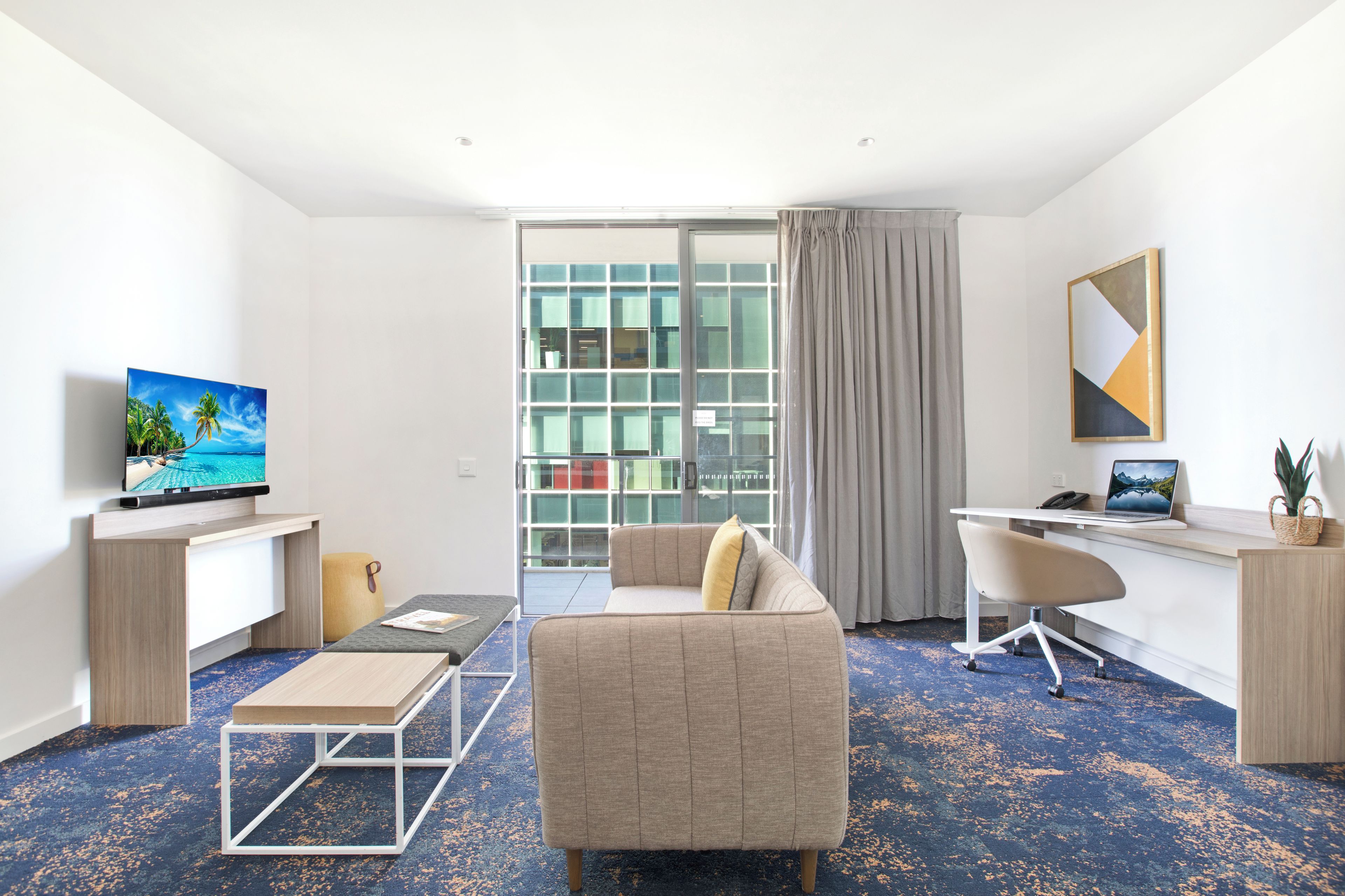 A modern hotel room with a sofa, a desk, a television, and a large window with curtains.