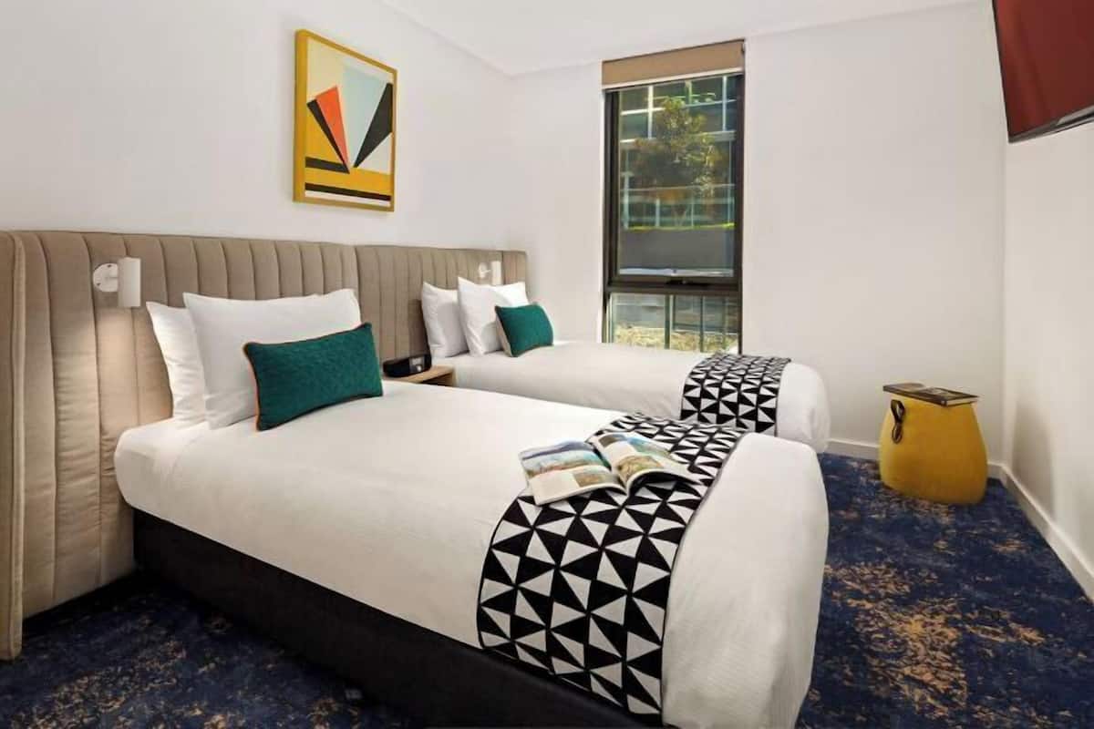 Habitación de hotel con dos camas, un ventanal grande, un cuadro en la pared y una pequeña mesa amarilla.