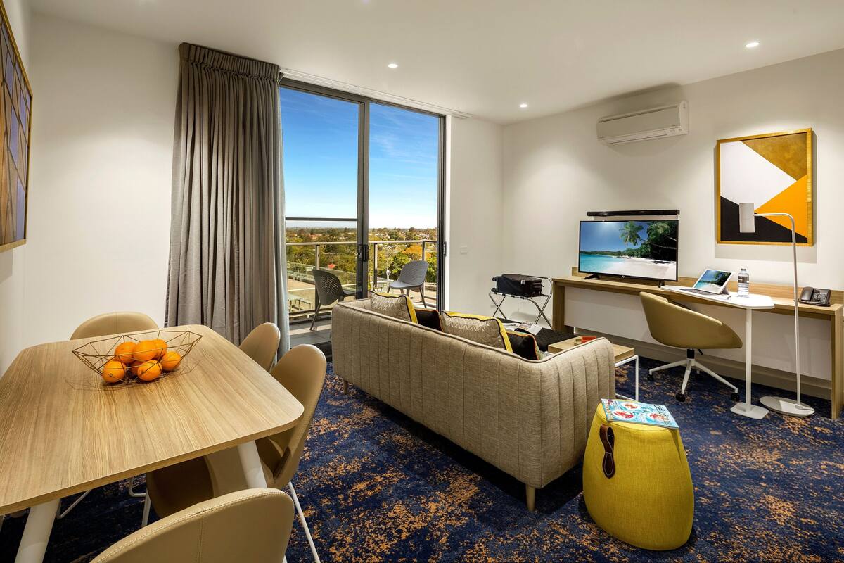 Habitación de hotel con un ventanal grande, un sofá, un escritorio con una computadora, un televisor y una mesa de comedor con naranjas.