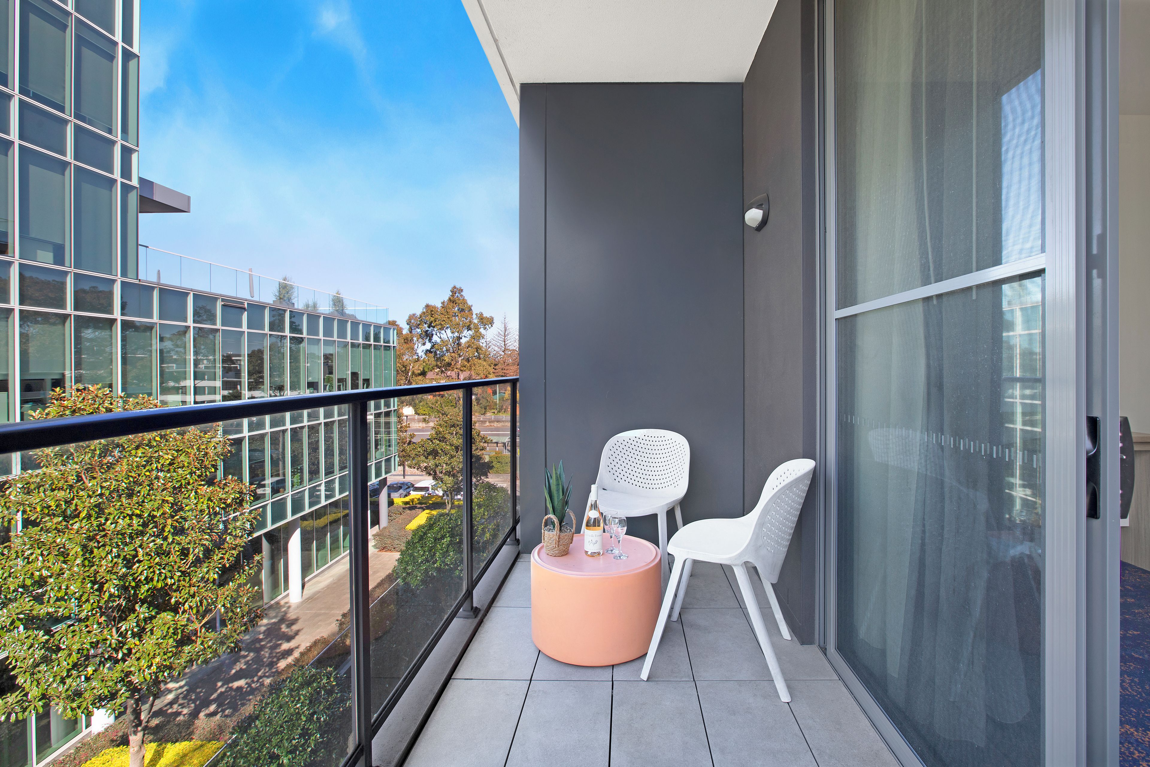 A balcony with a glass railing, a round table, two white chairs, and a view of a modern building and trees.