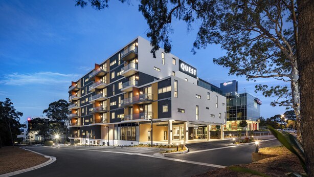 Un hotel de varios pisos con balcones, una entrada bien iluminada y un cartel visible que dice “QUEST”.