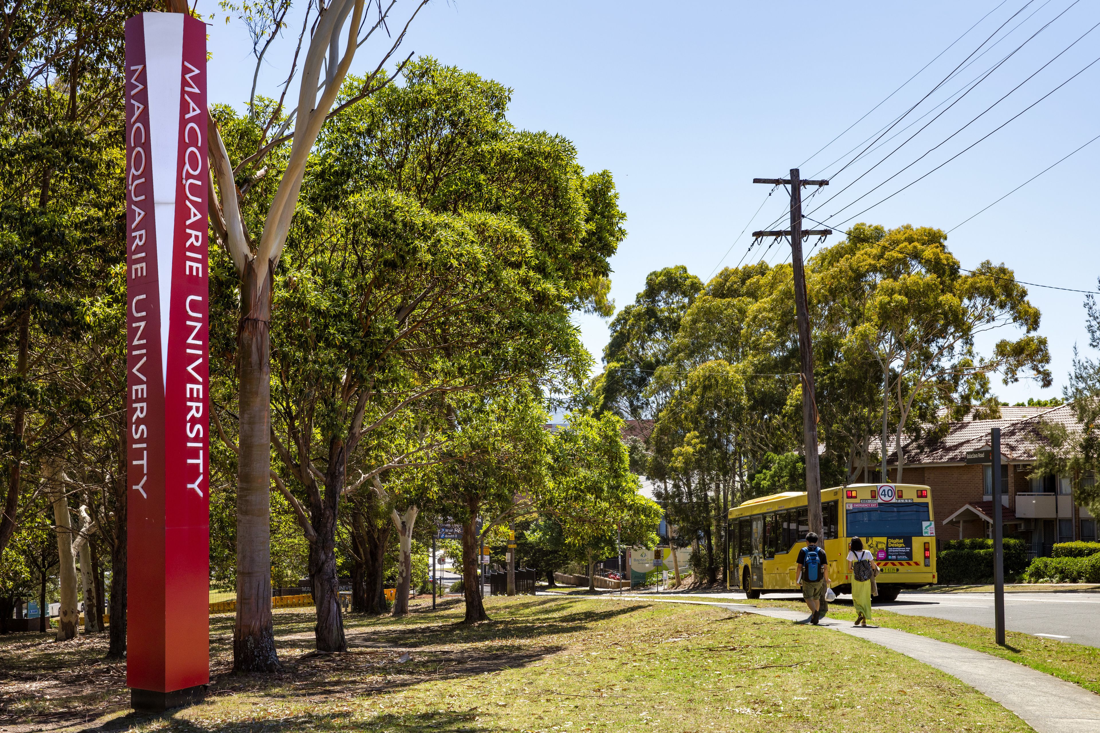 A university campus with a prominent red and white sign, lush green trees, and a yellow bus on the road.