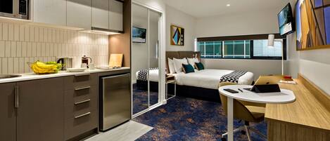A modern hotel room with a kitchenette, a bed with a view, and a desk with a laptop.