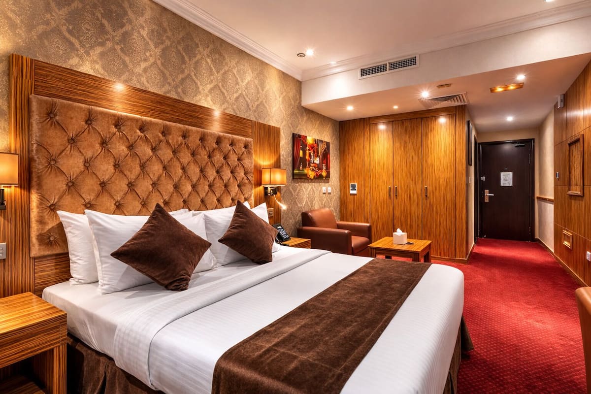 A hotel room with a large bed, a wooden headboard, a red carpet, a wooden wardrobe, and a seating area with a chair and table.