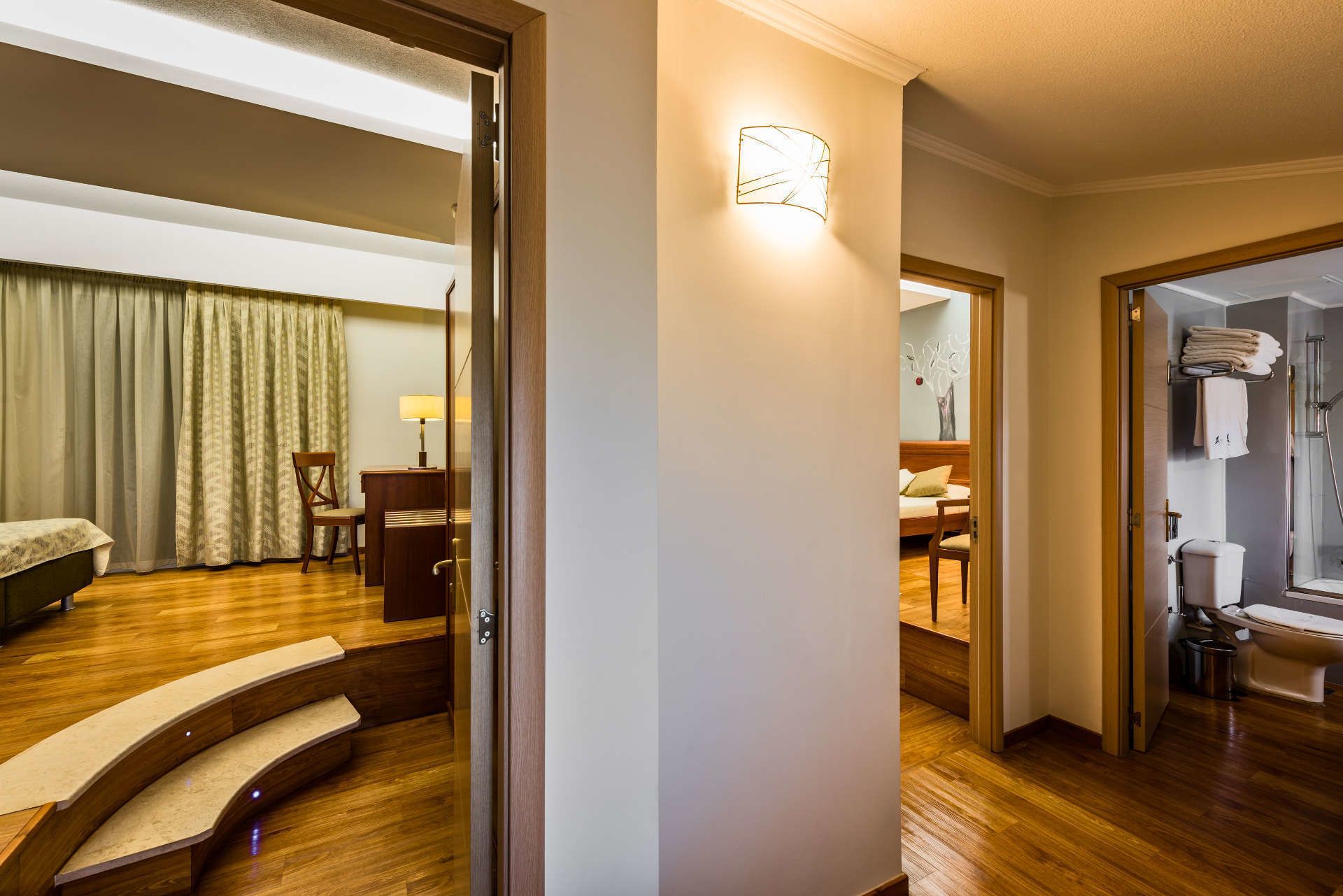 A hotel room with a wooden staircase, a desk with a lamp, a bed, and a bathroom with a shower and toilet.