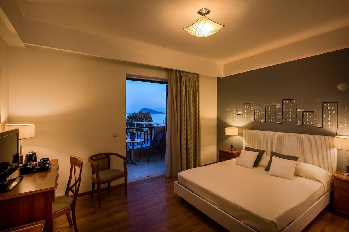 A hotel room with a bed, a desk, a chair, a balcony with a view of the sea, and a wall with framed pictures.