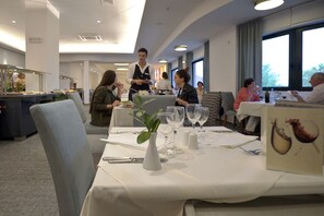 Uma sala de jantar com mesas postas para hóspedes, um garçom atendendo a uma mesa e uma área de buffet ao fundo.