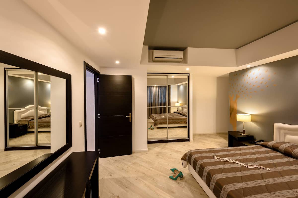A modern hotel room with a large bed, a mirror, a wardrobe, and a small table with a lamp.