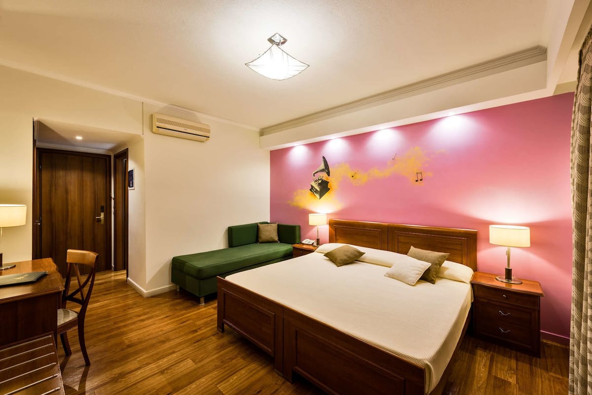 A hotel room with a large bed, a green sofa, a wooden desk with a chair, and a small table.