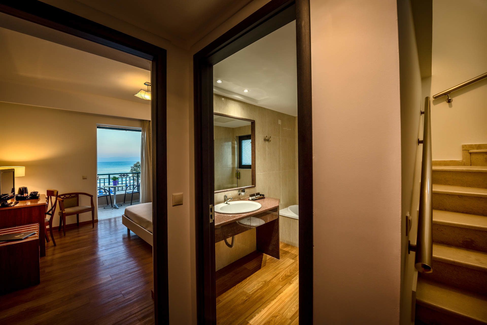 A hotel room with a balcony, a bed, a desk, a bathroom with a sink and mirror, and a staircase.