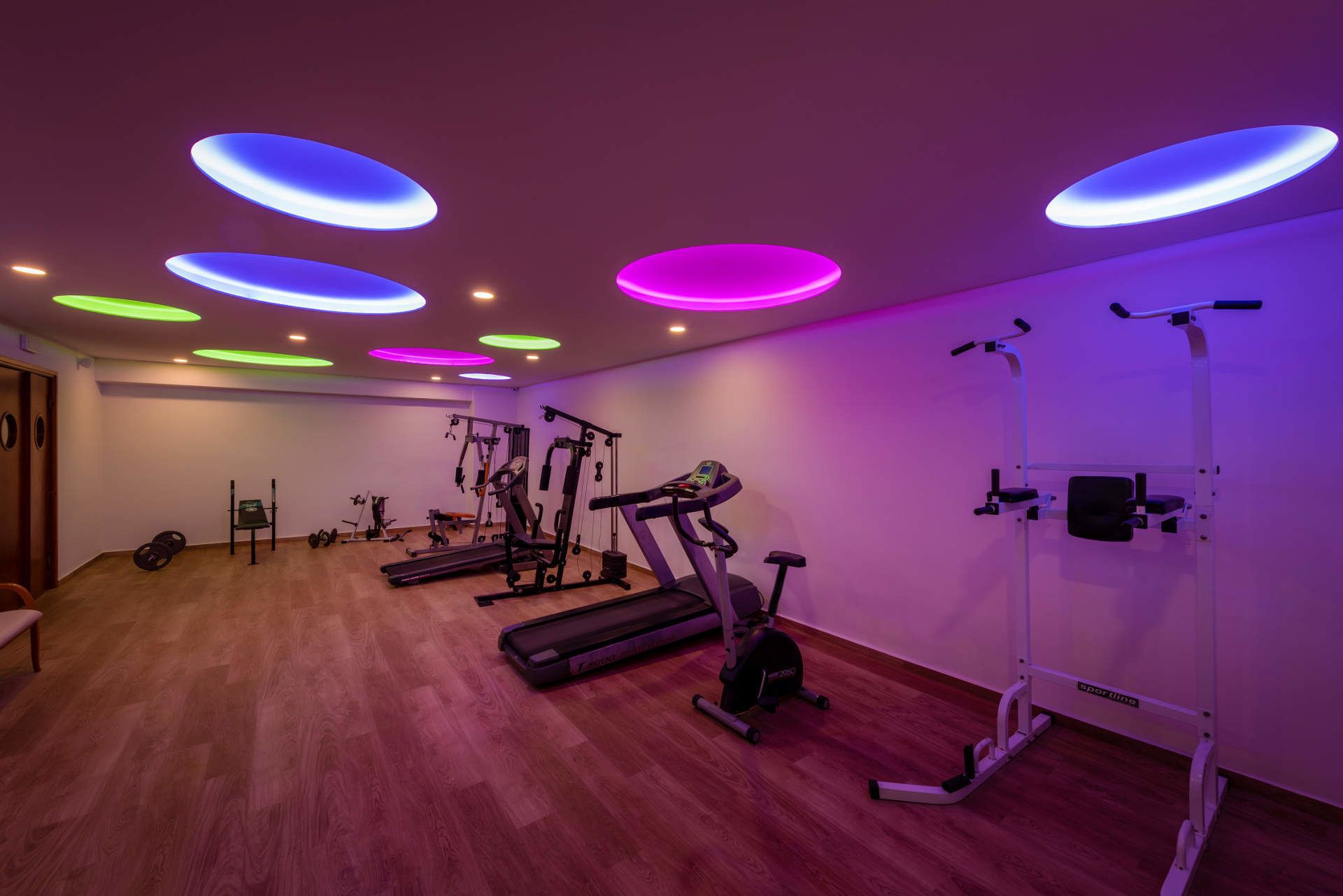 A gym with various exercise equipment, including treadmills and weight machines, under colorful ceiling lights.