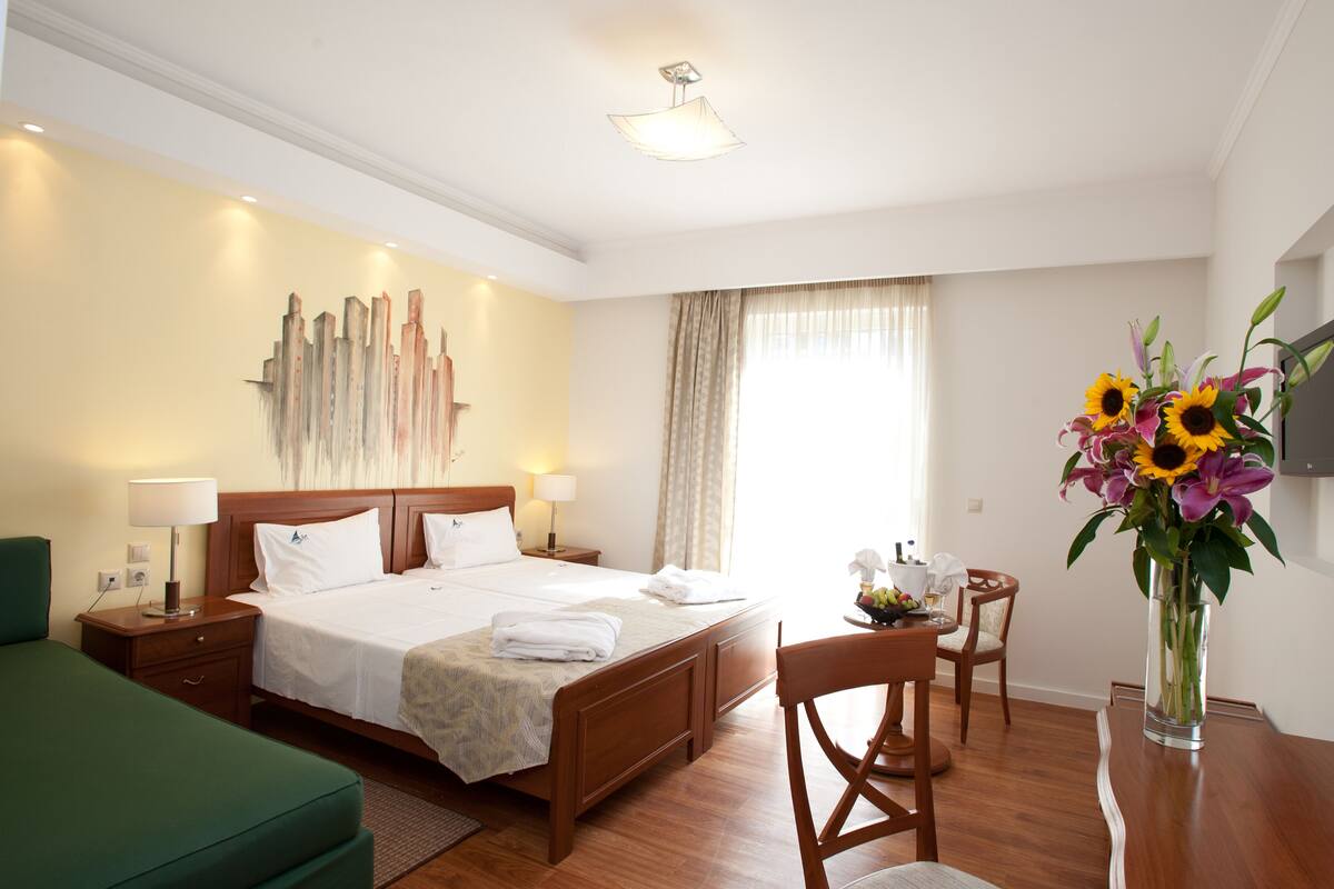 A hotel room with a large bed, a wooden nightstand, a green armchair, a dining table with chairs, a vase with flowers, and a wall-mounted TV.